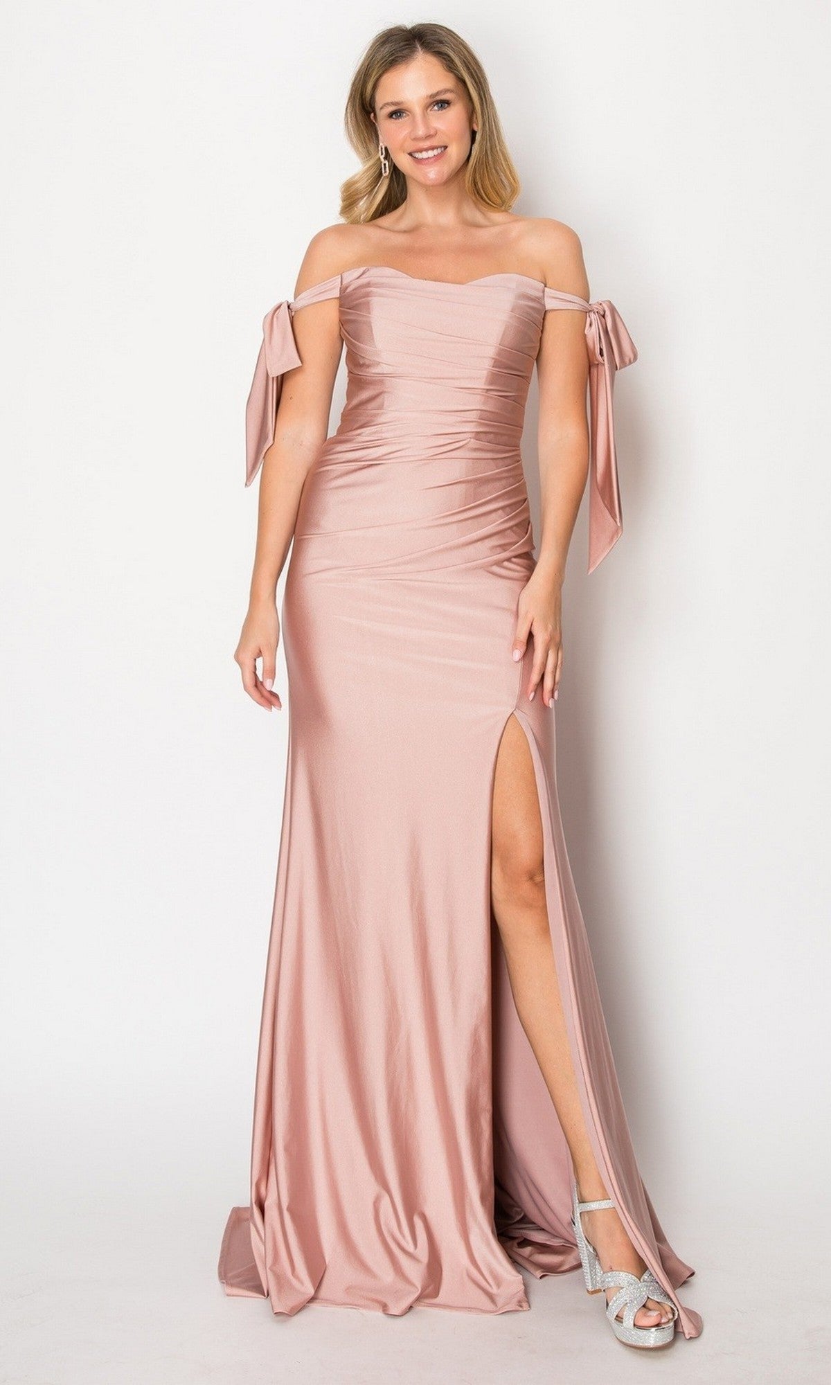 A woman wearing the Long Prom Dress: Chicas DM8010 in mocha, featuring an off-the-shoulder neckline and a thigh-high slit, stands smiling against a plain background, paired with silver heels.
